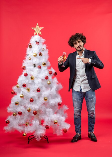 New year celebration with bearded young man pointing wine and standing near decorated white Christmas tree