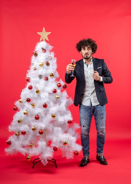 New year celebration with bearded young man with wine standing near decorated white Christmas tree on red