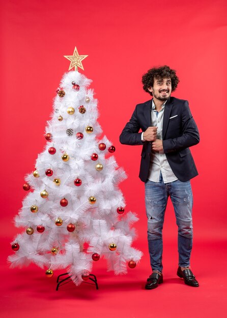 New year celebration with young man hiding something near decorated white Christmas tree on red