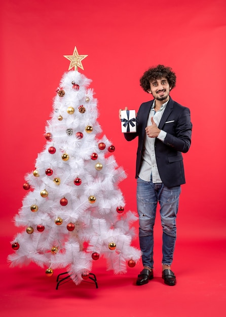 New year celebration with young man showing his gift making ok gesture near decorated white Christmas tree on red