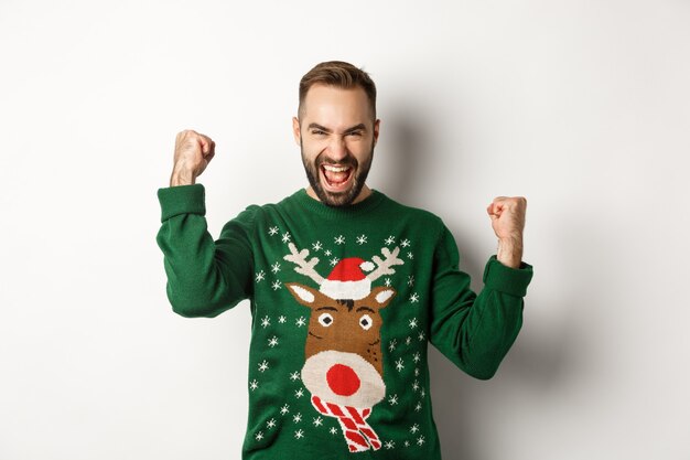 New Year, holidays and celebration. Excited bearded man in Christmas sweater, making fist pumps and shouting for joy, rejoicing and triumphing, white background.