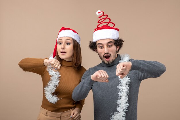 New year mood festive concept with excited cool satisfied lovely couple wearing red santa claus hats checking their time on gray