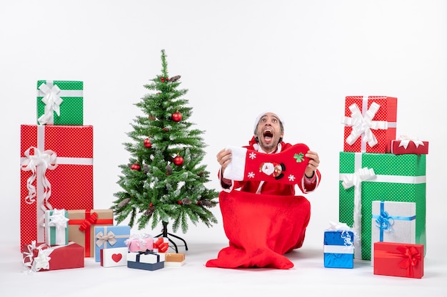 Free photo new year mood with funny positive surprised santa claus sitting on the ground and showing christmas sock near gifts and decorated xsmas tree on white background