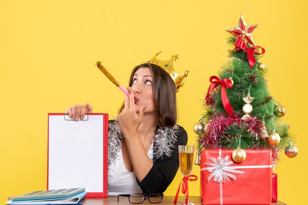 New year mood with thoughtful surprised charming lady in suit with crown holding document in the office on yellow isolated 