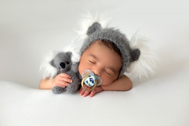 Newborn babyboy cute little resting baby with grey hat and grey toy bear in his hand and pacifier on his mouth on a white floor