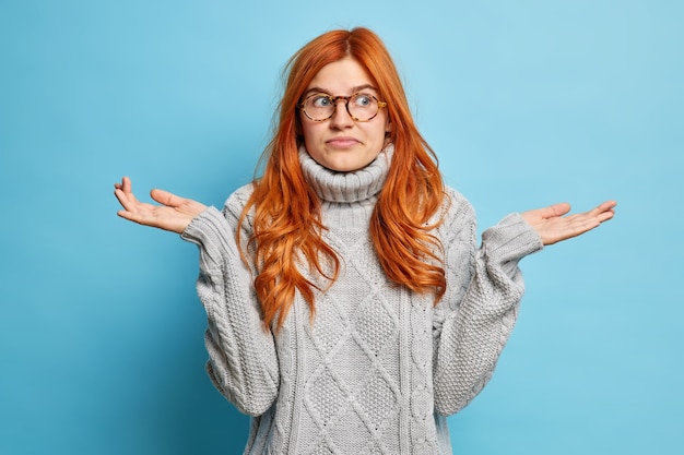 Free photo nice attractive doubtful woman shrugs shoulders doesnt know answer on difficult question doesnt care about your problems stands indifferent against blue wall has no knowledge shows uncertain gesture