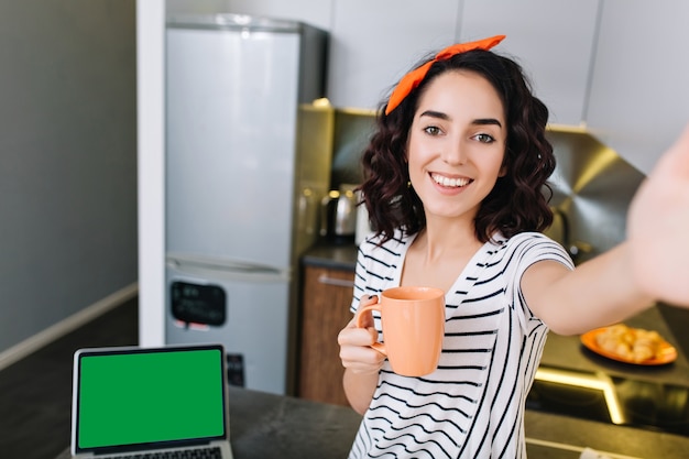 Nice beautiful selfie portrait of amazing joyful happy woman with cut curly brunette hair chilling in kitchen in modern apartment. Having fun, drinking tea