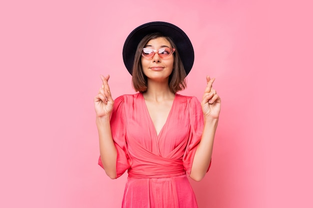 Nice cheerful woman wishes to do her best to keep her fingers crossed, has the best, wearing in dress  isolated on pink wall. People, body language concept. Copyspace.