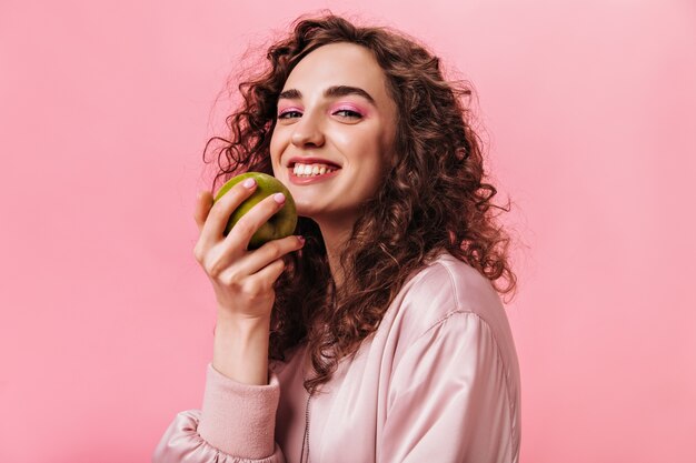 Nice woman in good mood holding green apple on pink background