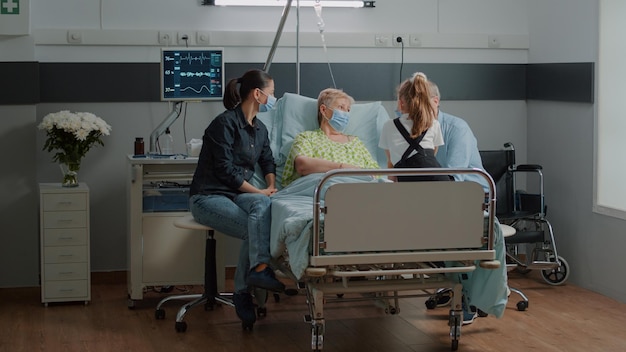 Free Photo niece visiting senior patient with face mask in hospital ward with mother and old man. sick woman enjoying visit from daughter and child, giving comfort to cure disease during pandemic.