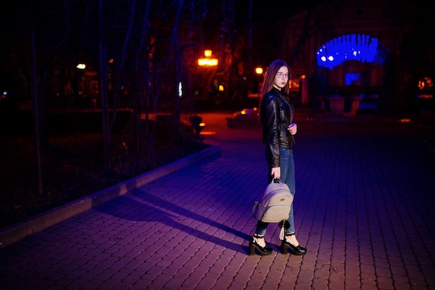 Free photo night portrait of girl model wear on glasses jeans and leather jacket with backpack in hands against blue lights garland of city street