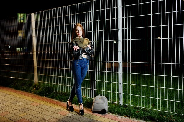 Night portrait of girl model wear on jeans and leather jacket against iron fence