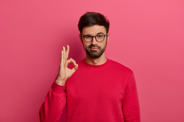 No problem concept. Bearded man makes okay gesture, has everything under control, all fine gesture, wears spectacles and jumper, poses against pink wall, says I got this, guarantees something