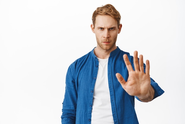 Free photo no stop serious and confident redhead man extend hand to block tell to stay away keep social distancing rejecting and forbidding something standing over white background