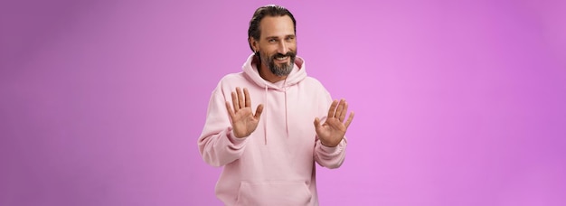 Free photo no thanks pass charming polite manly adult bearded man grey hair in pink hoodie apologizing refusal raise palms rejection gesture declining offer smiling say sorry not interested purple background