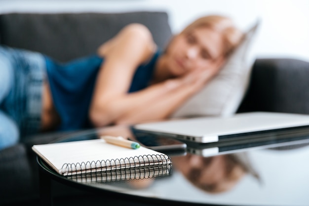 Notepad and laptop on table near woman sleeping on sofa