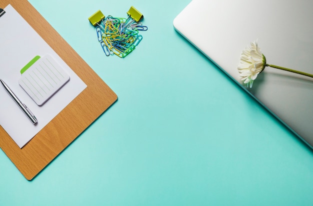 Notepad; pen; paper on clipboard with paperclips and laptop on green background