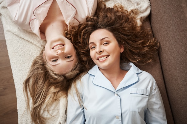 Nothing can be stronger that friendship. Two beautiful european women lying on sofa in comfortable nightwear, spending leisure together, smiling broadly, having girly talks