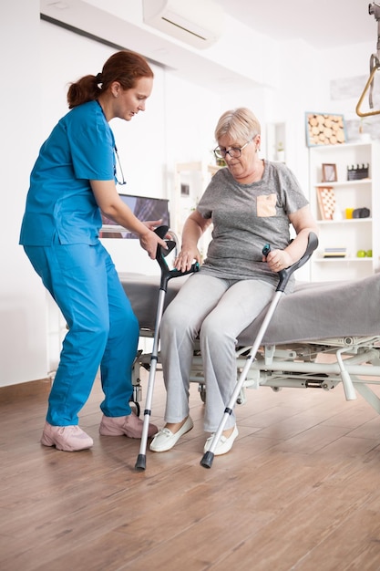 Nurse helping old woman to sit on bed in nursing home after walking with crutches.