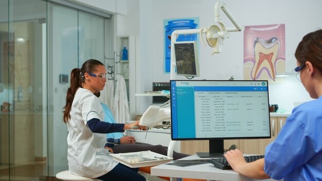 Nurse typing on computer, making appointments while dental specialist talking patient in stomatology chair before examination. Dentist and nurse working together in modern stomatological clinic