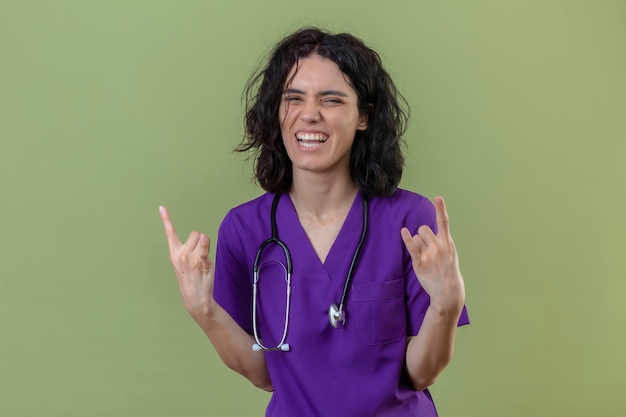 Free photo nurse wearing uniform and stethoscope smiling cheerful looking happy making rock symbols with fingers on isolated green