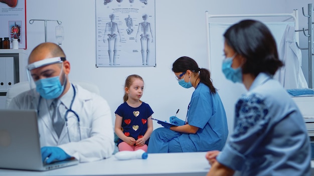 Free photo nurse with protection mask writing child data on clipboard during covid-19. physician specialist in medicine providing health care services consultation, treatment, examination in hospital cabinet