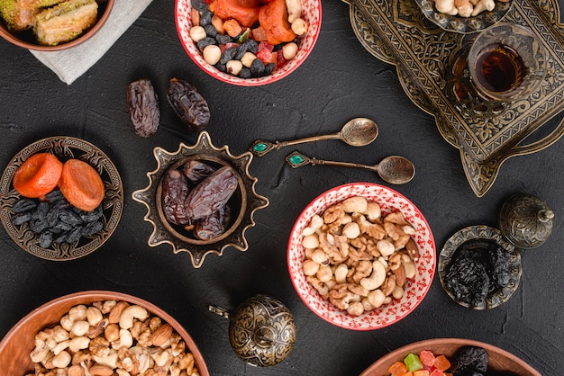 Free photo nuts; dried fruits and dates on metallic; spoons and ceramic bowl on black background