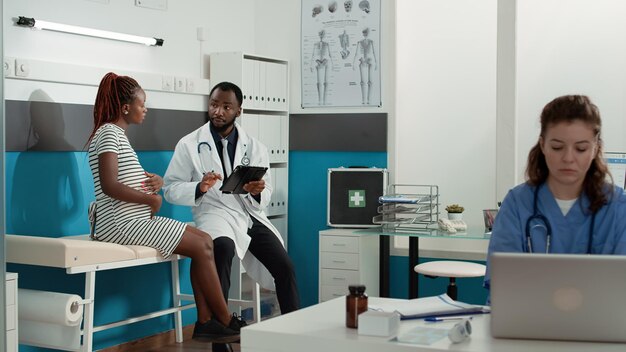 Obstetrician using digital tablet to consult patient expecting child at medical appointment. Male doctor taking notes at checkup visit examination, talking to woman with baby bump.