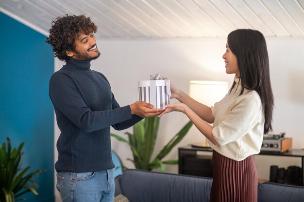 Occasion, gift. Profile of smiling young curly haired indian man and asian happy woman standing looking at each other touching gift box in lighted room