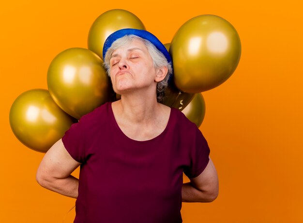 Offended elderly woman wearing party hat holds helium balloons behind with closed eyes on orange