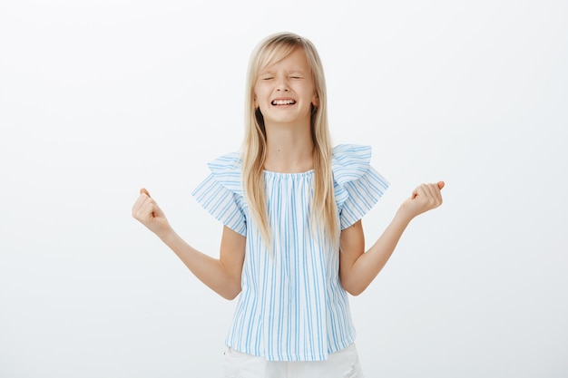 Offended little girl whining and crying, being disobedient. Displeased upset young daughter with blond hair, clenching spread fists and teeth, arguing wanting candy, standing over gray wall