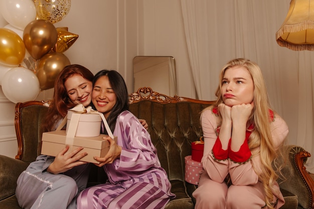 Offended young blonde woman in pajamas is sitting on sofa next to joyful girlfriends holding gift boxes Leisure and lifestyle concept