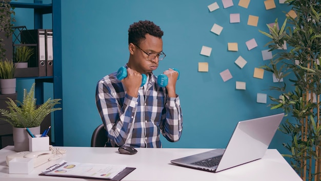 Office worker exercising with dumbbells to train muscles while he uses laptop at desk. Employee lifting weights and doing physical activity with fitness exercise, working on computer for business.