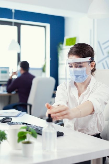 Free Photo office worker following safety precaution during global pandemic with coronavirus applying sanitizer. businesswoman in new normal workplace disinfecting while colleagues working in background.