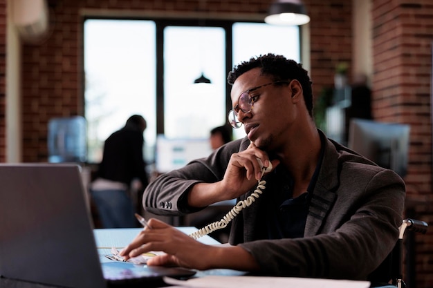 Office worker wheelchair user answering landline phone call, having remote conversation on telephone line in disability friendly office. Man with chronic impairment using telework chat line.