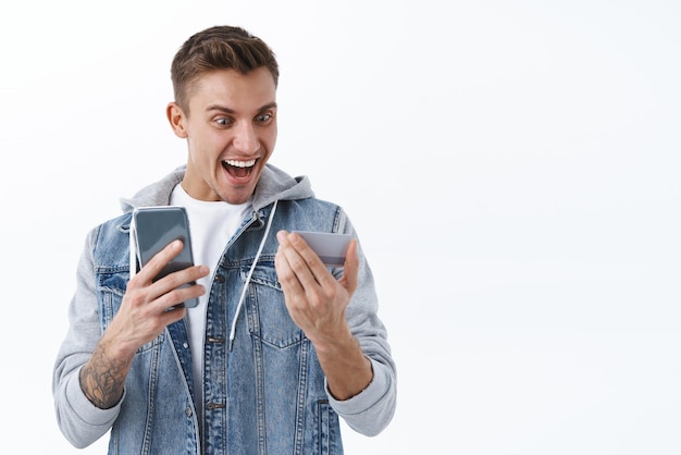 Oh gosh I won Celebration and prizes concept Portrait of young blond guy looking at credit card amused and excited smiling happy hurry up to order online shop with big discount hold smartphone