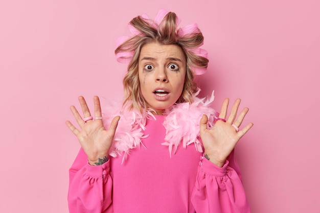 Free photo oh no its terrible. frightened emotional young woman stares with eyes full of fear keeps palms raised in defence tries to protect herself wears hair rollers and dress poses against pink background