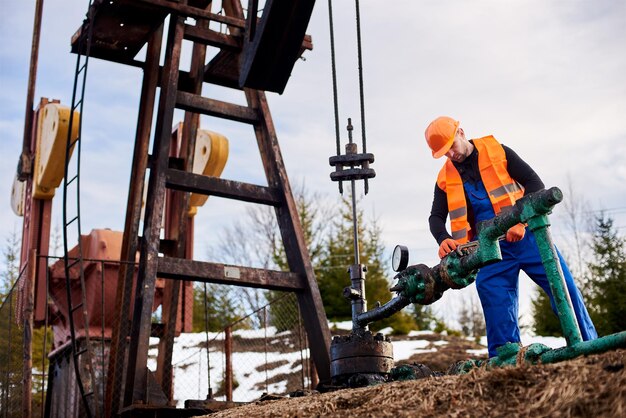 Oil worker in orange uniform and helmet working with a pipe wrench near an oil pump jack