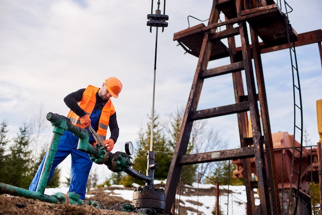 Oil worker in orange uniform and helmet working with a pipe wrench near an oil pump jack