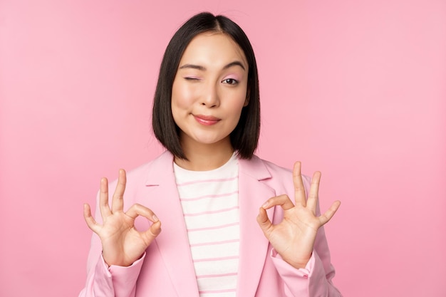 Okay excellent Businesswoman in corporate suit showing ok approval gesture recommending smth give positive feedback and smiling pleased posing over pink background