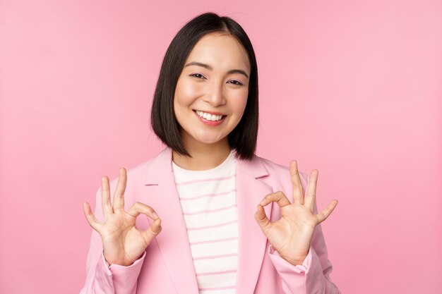 Okay excellent Businesswoman in corporate suit showing ok approval gesture recommending smth give positive feedback and smiling pleased posing over pink background