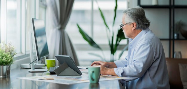 Old asian senior retired age working at home hand ise desktop and paper document at working desk in living room home interior backgroundasian male senior work at home