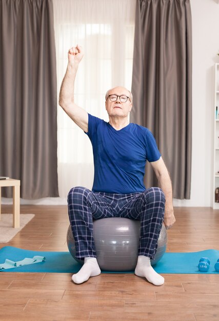 Old man training his arms sitting on stability ball in living room