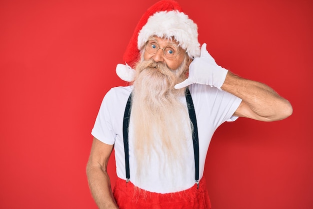 Old senior man with grey hair and long beard wearing white t-shirt and santa claus costume smiling doing phone gesture with hand and fingers like talking on the telephone. communicating concepts.