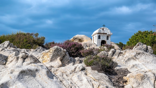 An old and small shrine located on rocks near the Aegean sea coast, bushes around, cloudy sky, Greece