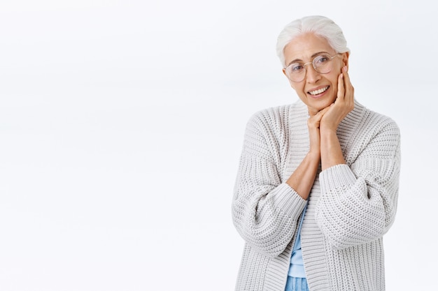 Old smiling, happy senior woman in glasses with grey hair, looking delighted and cheerful camera, grinning as touching face and contemplate grandchild playing