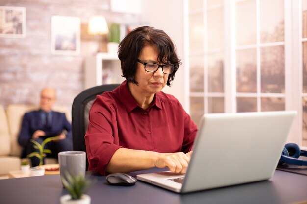 Old woman in her 60s using a modern laptop in her cozy house late in the evening