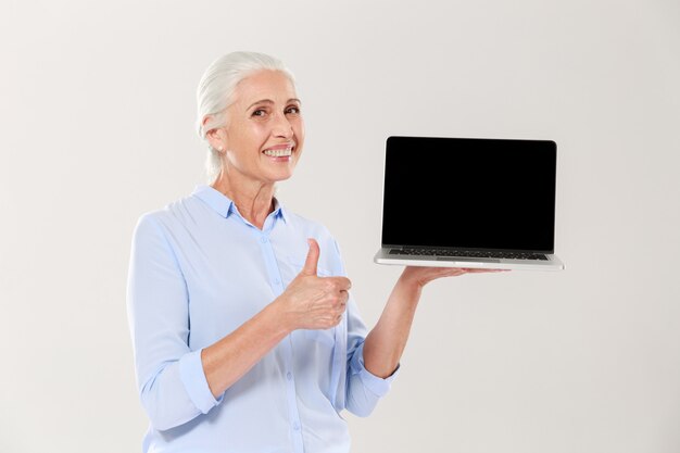 Old woman holding laptop and showing thumb up isolated