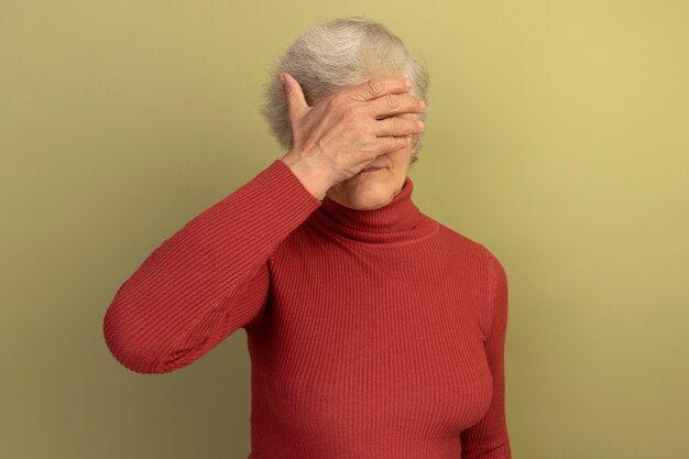 Old woman wearing red turtleneck sweater and sunglasses covering eyes with hand isolated on olive green wall with copy space