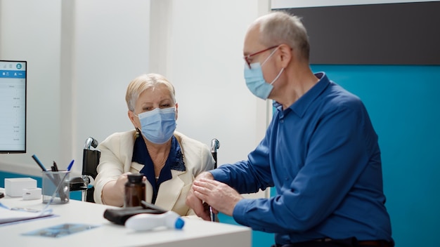 Old woman with chronic disability and her husband at appointment, attending checkup visit with medical specialist. Patient wheelchair user doing consultation during covid 19 pandemic.